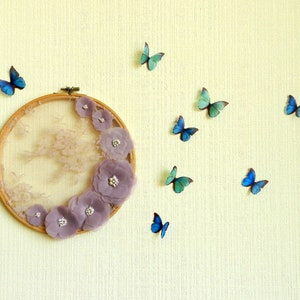 May include: A circular embroidery hoop with a lace background and purple fabric flowers. The hoop is decorated with blue butterflies on a light yellow wall.