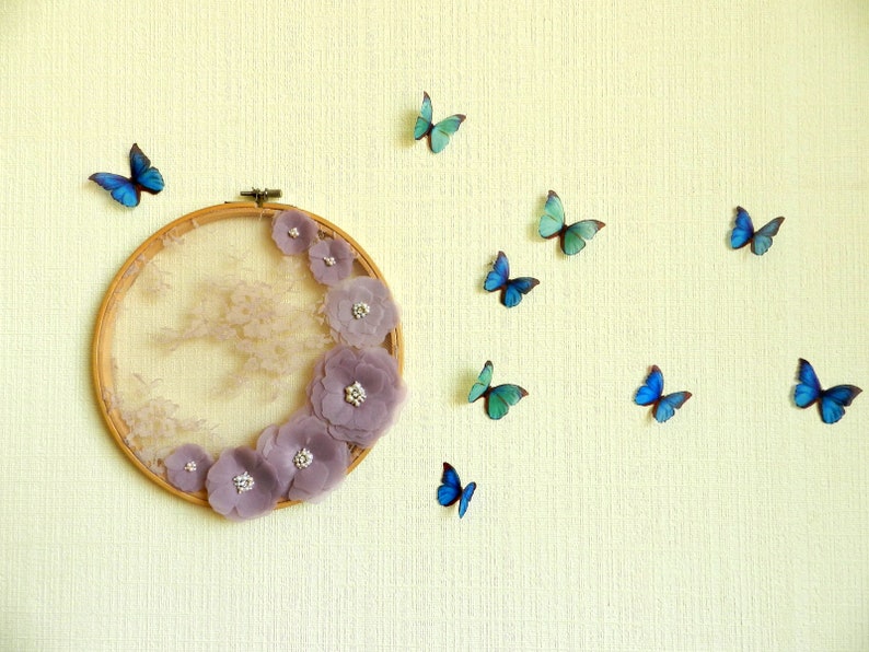 May include: A circular embroidery hoop with a lace background and purple fabric flowers. The hoop is decorated with blue butterflies on a light yellow wall.