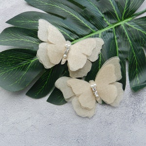 May include: Two delicate, beige butterfly hair clips with pearl accents. The clips are on a green leaf background.