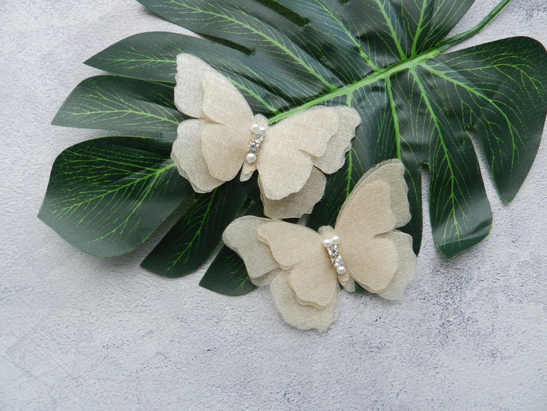 May include: Two delicate, beige butterfly hair clips with pearl accents. The clips are on a green leaf background.