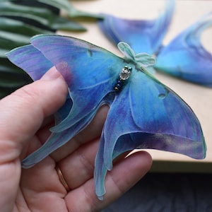 May include: A close-up of a blue and purple moth with delicate, sheer wings. The moth has a small, jeweled embellishment on its body. The wings have a gradient effect, transitioning from blue to purple.