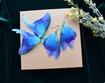 Puede incluir: Un conjunto de pendientes y un broche con forma de alas de mariposa azules. Las alas tienen un degradado de azul y morado, con ganchos plateados en los pendientes. El broche tiene un centro con cuentas. Los artículos se muestran en una caja beige.