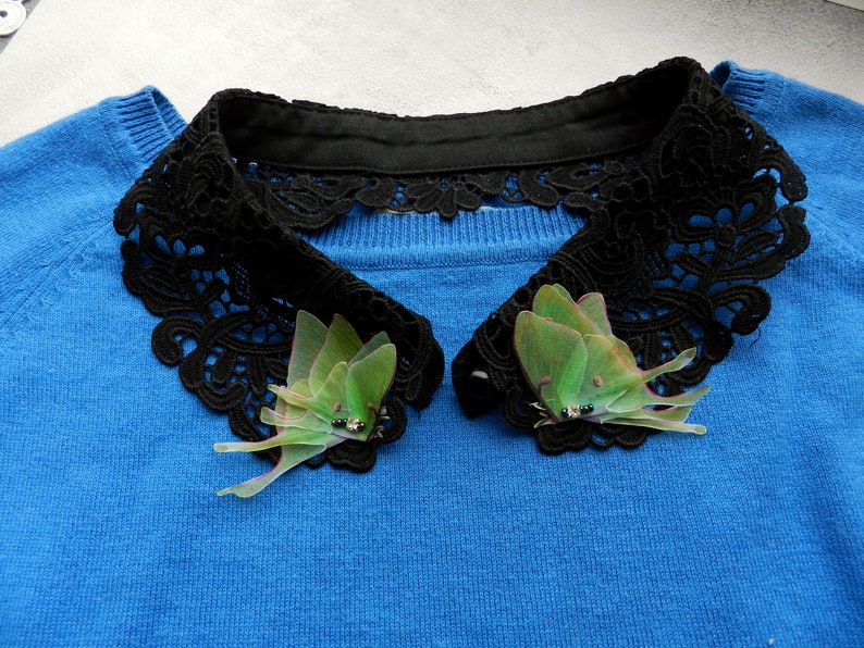K&ouml;nnte beinhalten: Ein blauer Pullover mit einem schwarzen Spitzenkragen und zwei gr&uuml;nen Mottenbroschen.