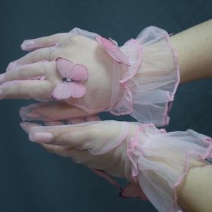 May include: Sheer pink gloves with delicate butterfly accents. The gloves feature a ruffled trim in a matching pink hue. The butterflies are also pink and add a whimsical touch to the design. A feminine accessory.