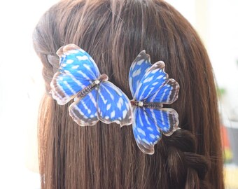 May include: Two butterfly hair clips with blue and white patterned wings are attached to brown hair. The butterfly wings have a detailed design with brown edges. The hair is styled in a braid.
