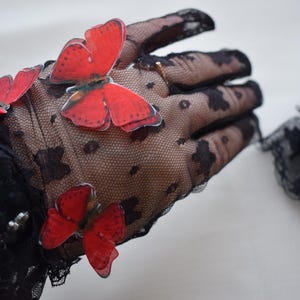 May include: Black lace gloves adorned with vibrant red butterfly appliqu&eacute;s. The gloves feature a delicate, dotted pattern and scalloped edges, creating an elegant and whimsical accessory. Perfect for special occasions or adding a touch of flair.