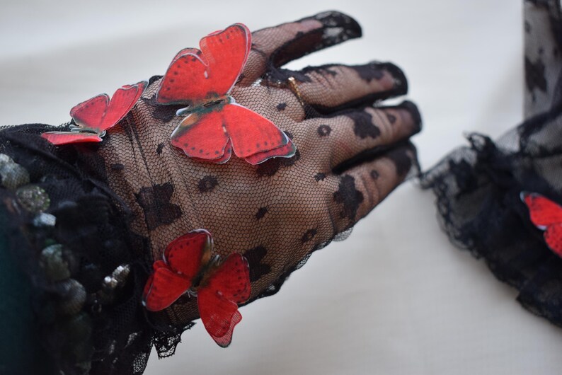 May include: Black lace gloves adorned with vibrant red butterfly appliqu&eacute;s. The gloves feature a delicate, dotted pattern and scalloped edges, creating an elegant and whimsical accessory. Perfect for special occasions or adding a touch of flair.