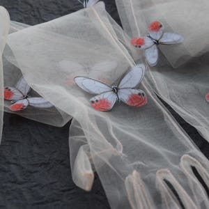May include: Close-up of sheer, off-white gloves adorned with butterfly appliqu&eacute;s. The butterflies have pale blue wings with red and white accents. The gloves are arranged on a dark, textured surface, creating a delicate and whimsical aesthetic.
