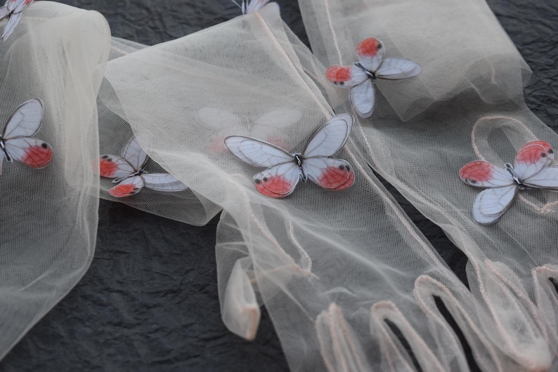 May include: Close-up of sheer, off-white gloves adorned with butterfly appliqu&eacute;s. The butterflies have pale blue wings with red and white accents. The gloves are arranged on a dark, textured surface, creating a delicate and whimsical aesthetic.