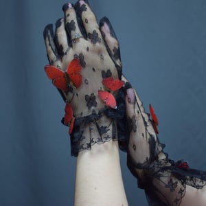 May include: Black lace gloves adorned with red butterfly embellishments. The gloves feature a floral pattern and a ruffled lace trim at the wrist. The butterflies add a whimsical touch to the elegant design.