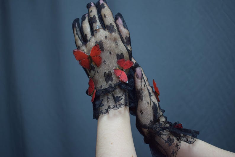 May include: Black lace gloves adorned with red butterfly embellishments. The gloves feature a floral pattern and a ruffled lace trim at the wrist. The butterflies add a whimsical touch to the elegant design.