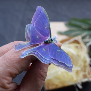 May include: A purple and green iridescent butterfly with a clear rhinestone on its thorax. The butterfly is made of fabric and is being held by a hand.