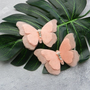 May include: Two peach-colored fabric butterfly hair clips with a pearl-like embellishment in the center of each butterfly.