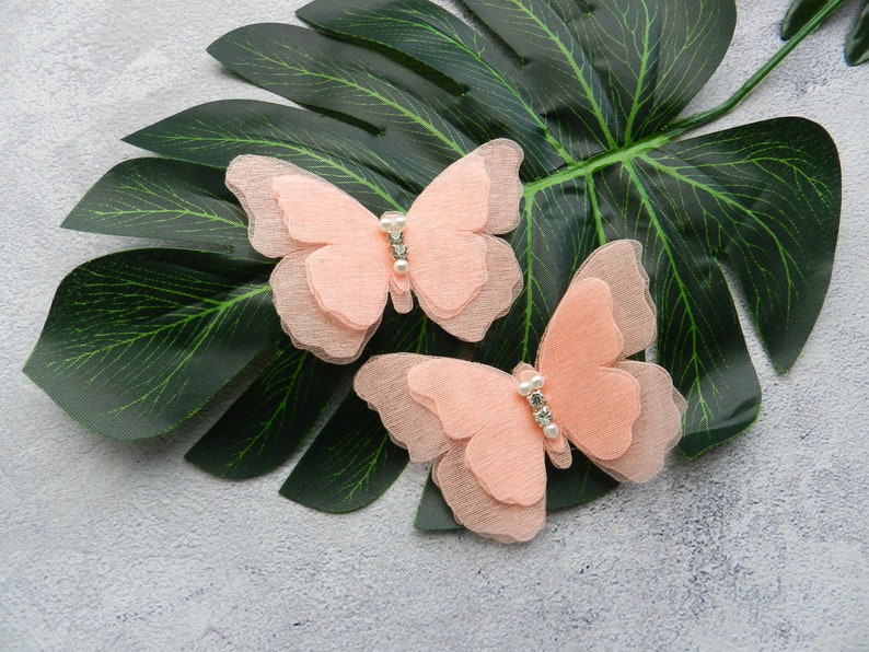 May include: Two peach-colored fabric butterfly hair clips with a pearl-like embellishment in the center of each butterfly.
