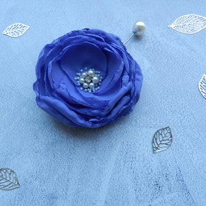 May include: A blue fabric flower brooch with a pearl center and a silver pin. The flower is surrounded by silver leaves.