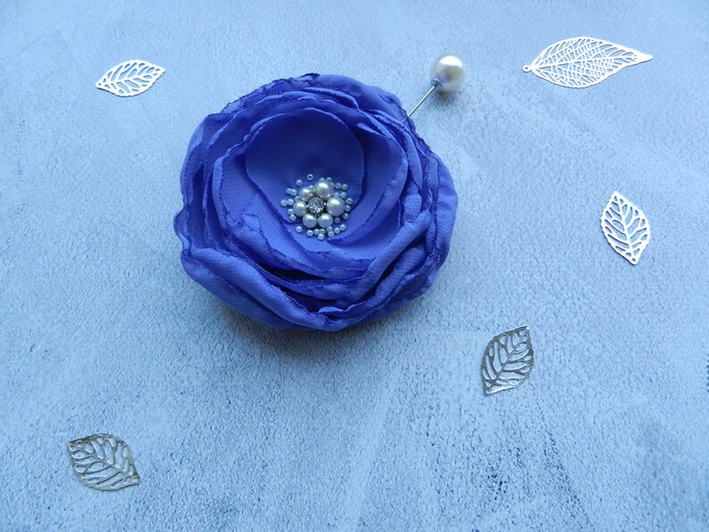 May include: A blue fabric flower brooch with a pearl center and a silver pin. The flower is surrounded by silver leaves.
