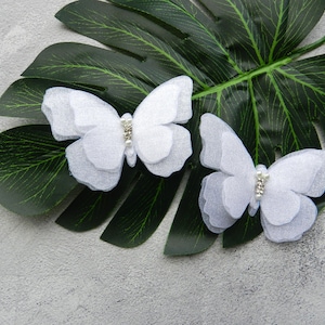 May include: Two white fabric butterfly hair clips with rhinestone accents on a green leaf background.
