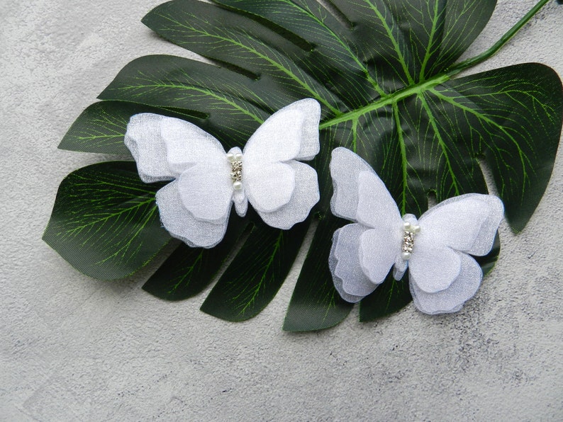 May include: Two white fabric butterfly hair clips with rhinestone accents on a green leaf background.