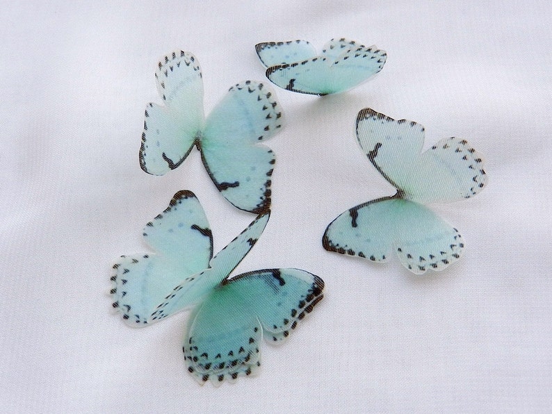 May include: Five light blue butterflies with black markings on a white background. The butterflies are made of a delicate material and have a translucent appearance.