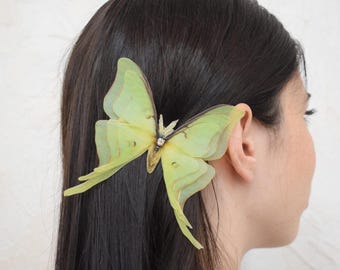 May include: A pale green butterfly hair clip with delicate, layered wings and a sparkling center. The accessory is attached to dark hair, positioned near the ear. The butterfly design adds a whimsical touch to the hairstyle.