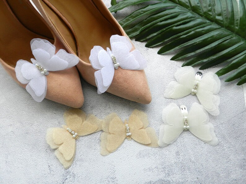 May include: A pair of beige shoes with white and beige butterfly embellishments. The butterflies are made of tulle and have pearl accents.