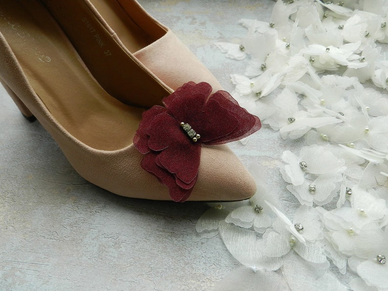 May include: A pair of nude-colored high heels with a burgundy butterfly embellishment on the toe. The butterfly has a rhinestone in the center.