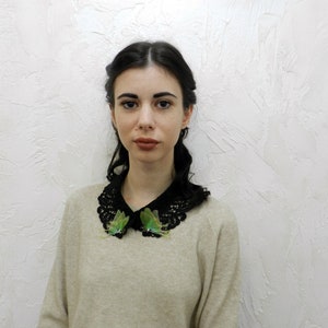 K&ouml;nnte beinhalten: Eine Frau mit dunklen Haaren tr&auml;gt einen beigefarbenen Pullover mit einem schwarzen Spitzenkragen. Der Kragen ist mit zwei gr&uuml;nen Schmetterlingsverzierungen verziert.