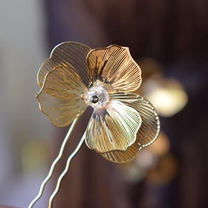 May include: A gold-colored floral hair pin with a delicate, layered petal design. The flower features a clear crystal center and is attached to a thin, wavy gold pin. The background is blurred, highlighting the intricate details of the hair accessory.