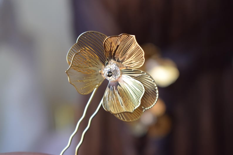 May include: A gold-colored floral hair pin with a delicate, layered petal design. The flower features a clear crystal center and is attached to a thin, wavy gold pin. The background is blurred, highlighting the intricate details of the hair accessory.