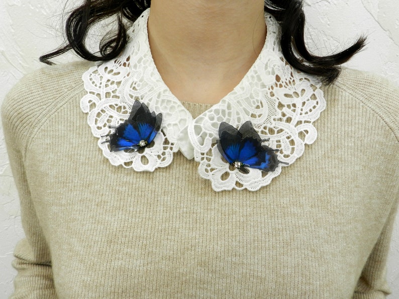 K&ouml;nnte beinhalten: Ein wei&szlig;er Spitzenkragen mit zwei blauen Schmetterlingsbroschen. Die Schmetterlinge haben schwarze Fl&uuml;gel und einen blauen K&ouml;rper mit einem silbernen Akzent.