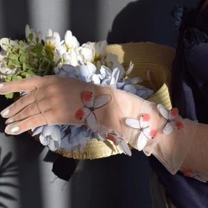 May include: Sheer, light-colored gloves adorned with butterfly appliqu&eacute;s. The gloves are worn while holding a straw hat filled with white and blue flowers. The butterflies have red and white details. The image is well-lit.