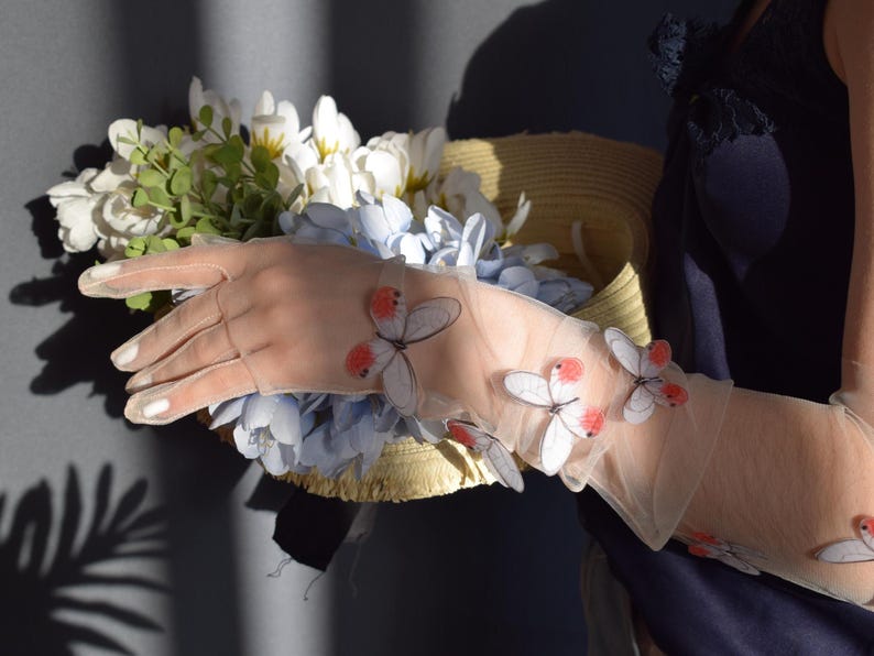 May include: Sheer, light-colored gloves adorned with butterfly appliqu&eacute;s. The gloves are worn while holding a straw hat filled with white and blue flowers. The butterflies have red and white details. The image is well-lit.