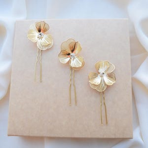 May include: Three gold-colored hair pins with flower-shaped designs. Each pin features a delicate, openwork flower with a small crystal center. The pins are arranged on a light brown surface, with a soft, white fabric in the background.