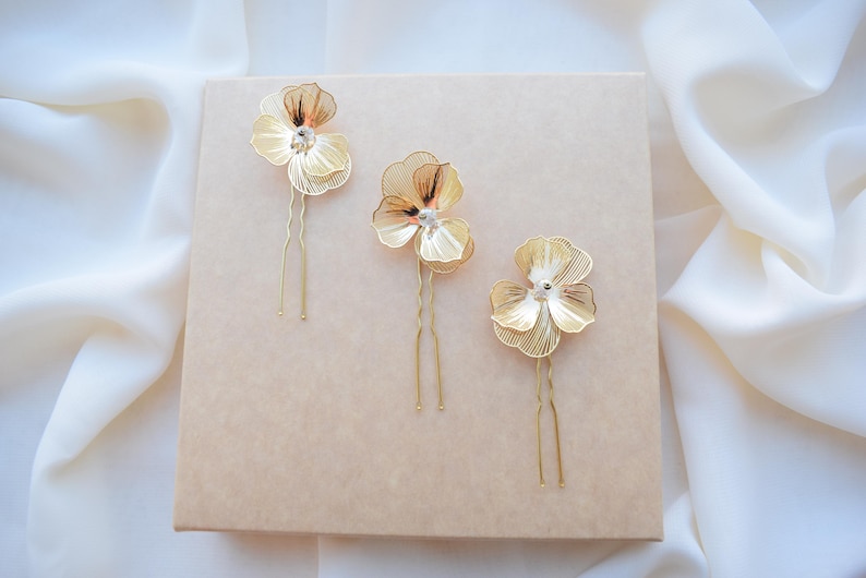 May include: Three gold-colored hair pins with flower-shaped designs. Each pin features a delicate, openwork flower with a small crystal center. The pins are arranged on a light brown surface, with a soft, white fabric in the background.