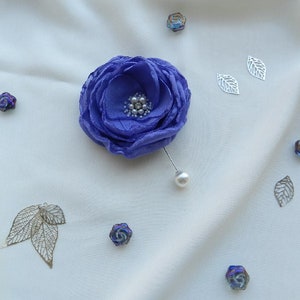 May include: A purple fabric flower brooch with a pearl accent. The brooch is pinned to a white fabric background.