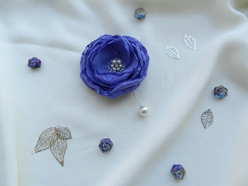 May include: A purple fabric flower brooch with a pearl accent. The brooch is pinned to a white fabric background.