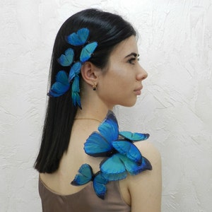 Pode incluir: Uma mulher com cabelo comprido e preto adornado com grampos de cabelo de borboleta azul. Ela está vestindo um top marrom sem mangas e tem asas de borboleta azuis nos ombros.