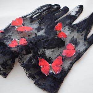May include: A pair of black lace gloves adorned with red butterfly appliqu&eacute;s. The gloves have a delicate, sheer appearance, with the butterflies adding a pop of color and a whimsical touch. The gloves are likely for a special occasion.