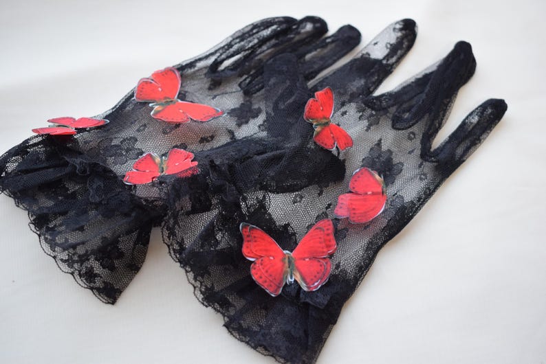 May include: A pair of black lace gloves adorned with red butterfly appliqu&eacute;s. The gloves have a delicate, sheer appearance, with the butterflies adding a pop of color and a whimsical touch. The gloves are likely for a special occasion.