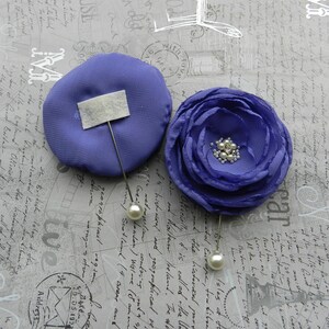 May include: Two purple fabric flower brooches with pearl accents. The brooches are pinned to a gray background with writing.