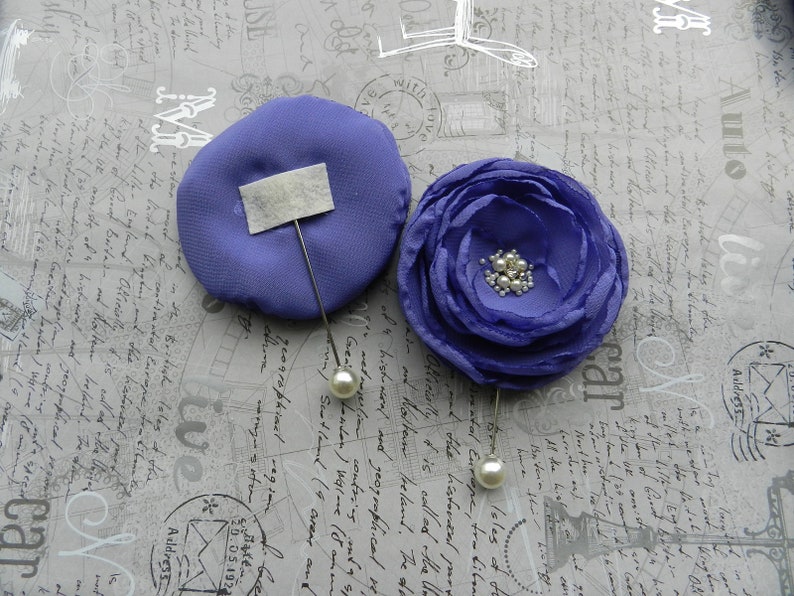May include: Two purple fabric flower brooches with pearl accents. The brooches are pinned to a gray background with writing.