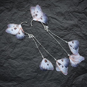 Puede incluir: Dos pares de pendientes con forma de alas de mariposa. Las alas son de color azul pálido con marcas azules oscuras y marrones. Los pendientes tienen cadenas y ganchos para las orejas de color plateado. El fondo es una superficie negra texturizada.