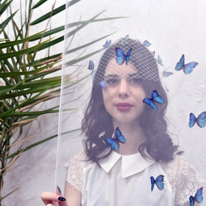 May include: A woman wearing a white veil with blue butterfly appliques. The veil is sheer and covers her face. She is wearing a white lace top and has dark hair.