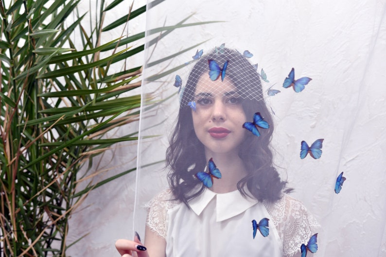 May include: A woman wearing a white veil with blue butterfly appliques. The veil is sheer and covers her face. She is wearing a white lace top and has dark hair.