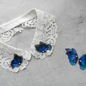 K&ouml;nnte beinhalten: Ein wei&szlig;er Spitzenkragen mit zwei blauen Schmetterlingsverzierungen. Der Kragen ist aus zarter Spitze gefertigt und hat einen gewellten Rand. Die Schmetterlinge sind aus Stoff gefertigt und haben ein realistisches Aussehen.