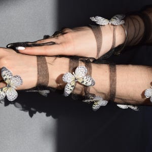 May include: Black sheer gloves and armbands adorned with delicate butterfly embellishments. The butterflies feature a detailed wing pattern in shades of white, gray, and black. The gloves and armbands are layered over the skin.
