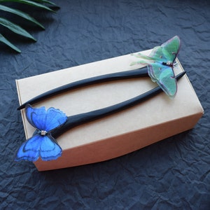 May include: Two black wooden hair sticks with butterfly embellishments. One stick has a blue butterfly with a rhinestone centre, the other has a green butterfly with a black and white pattern.