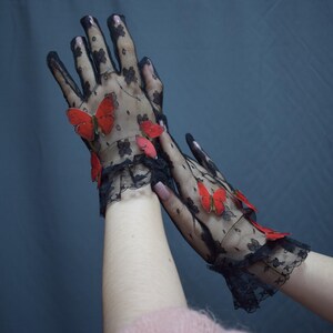 May include: Black lace gloves adorned with red butterfly accents. The gloves feature a delicate floral pattern and a ruffled trim at the wrist, creating an elegant and whimsical accessory.