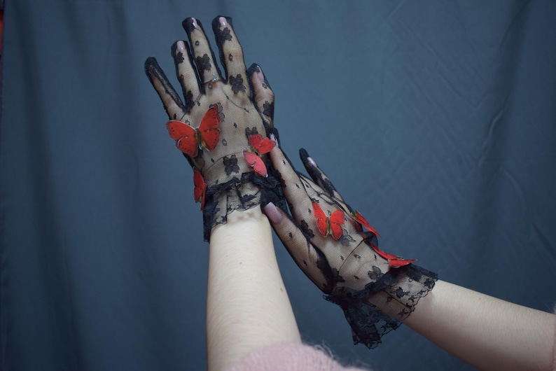May include: Black lace gloves adorned with red butterfly accents. The gloves feature a delicate floral pattern and a ruffled trim at the wrist, creating an elegant and whimsical accessory.