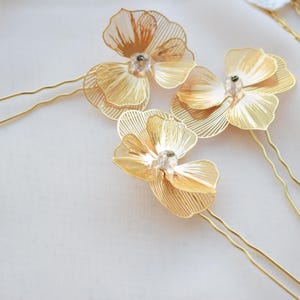May include: Gold-toned metal hair pins with delicate, openwork flower designs. Each flower features a central crystal accent. The pins are arranged on a soft, white surface, suggesting a bridal or special occasion theme.